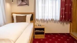 Ein Hotelzimmer mit einem Bett, einem Nachttisch mit Buch und Brille, einer Wandleuchte, einem Fenster mit durchsichtigen Vorhängen und einem roten Teppich mit Muster.