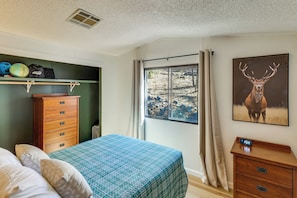 A bedroom with a bed, a dresser, a shelf with helmets and a bag, a window with a view of the outdoors, and a deer painting on the wall.