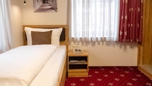 A hotel room with a bed, a nightstand with a book and glasses, a wall-mounted light, a window with sheer curtains, and a red patterned carpet. - Comfortable single room 7 in the historic center of Heidenheim (Heidenheim)