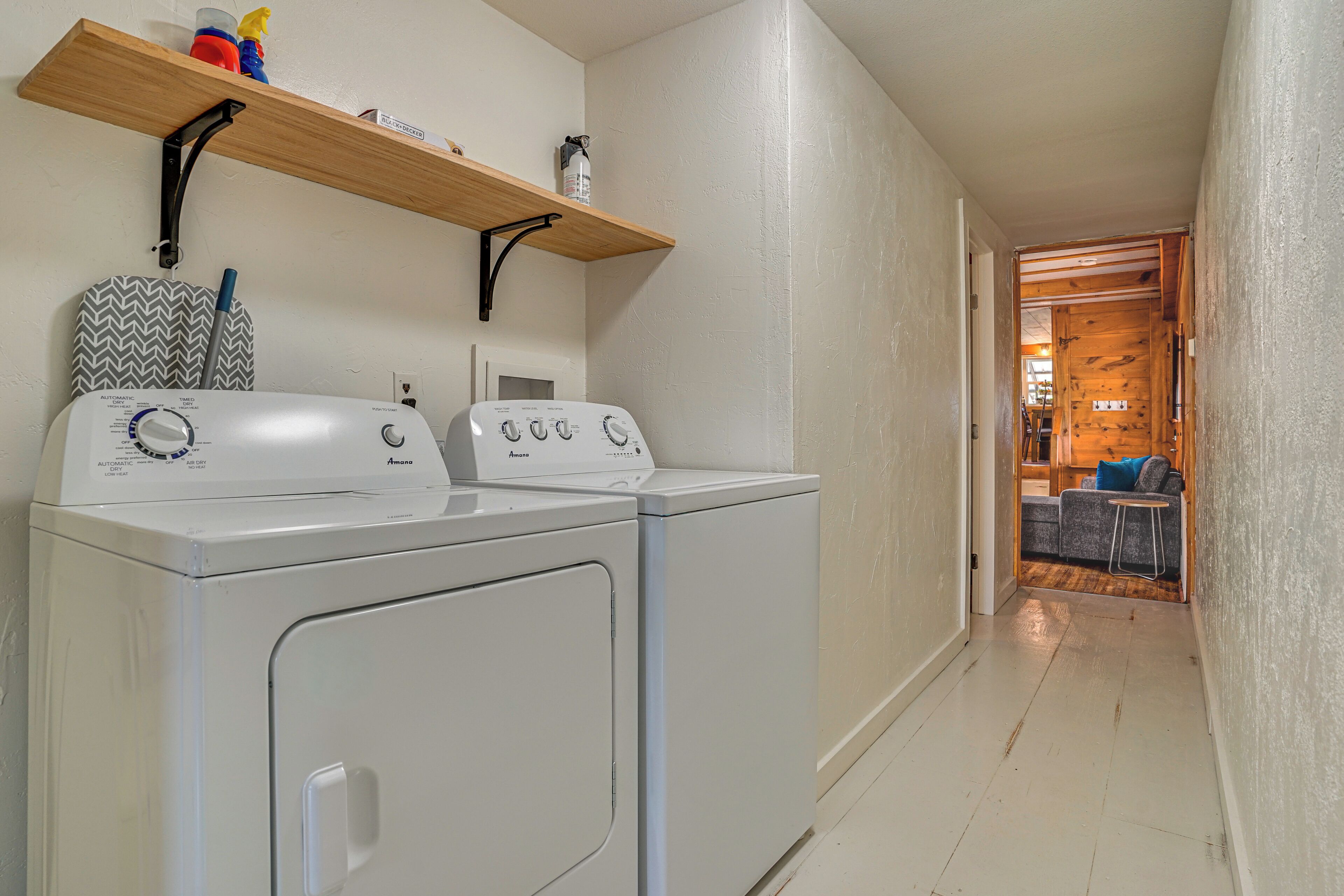 A laundry area with two front-loading washers and dryers, a wooden shelf above, and a hallway leading to a living area.