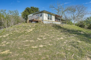 A single-story house with a deck, surrounded by a grassy hill and trees. - Serene Hilltop Hideaway: 7 Mi to Weatherford! (Weatherford)