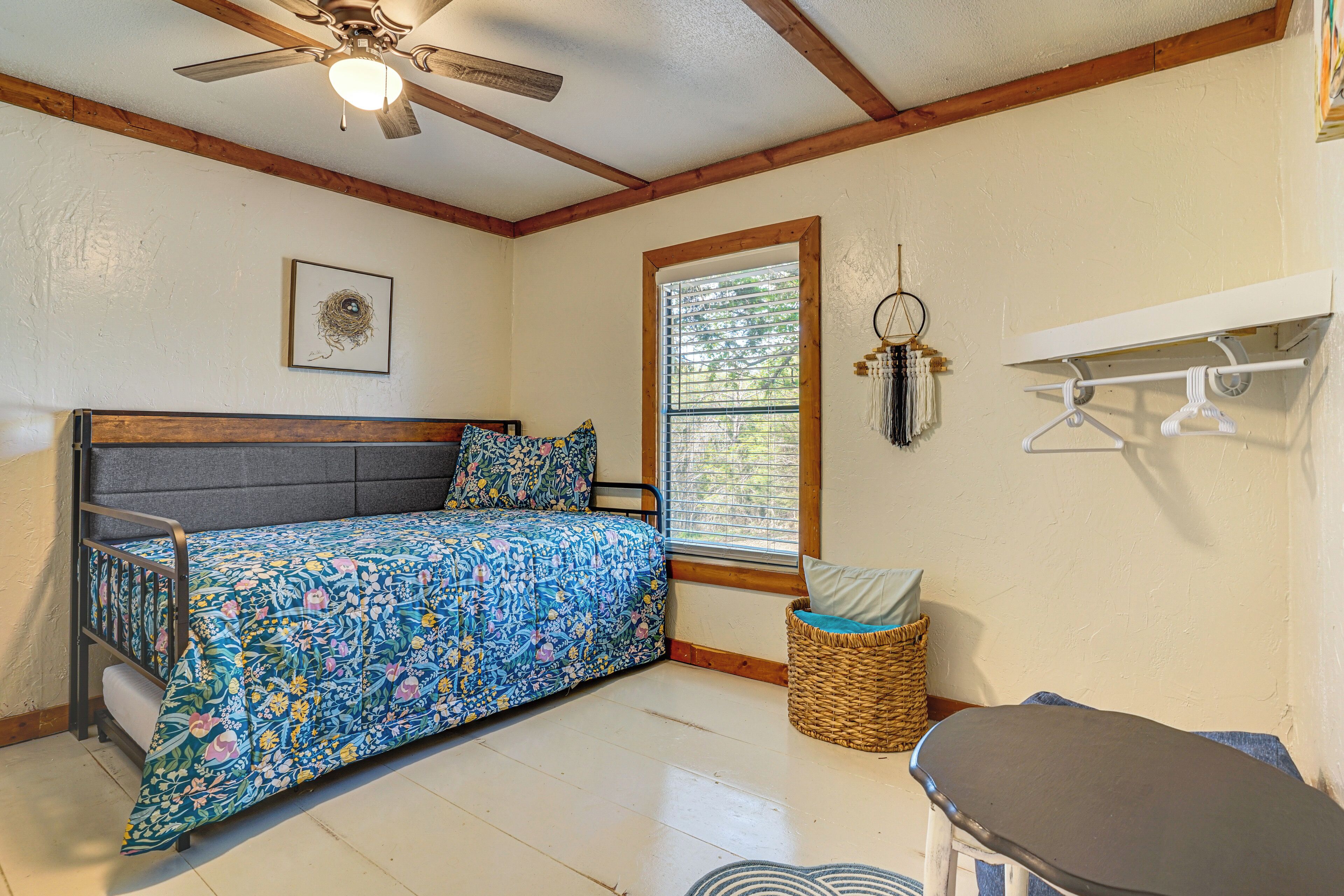 A room with a bed, a ceiling fan, a window, a wall-mounted rack, and a small table.