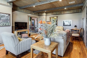 A spacious living room with a fireplace, wooden ceiling, and a large window.