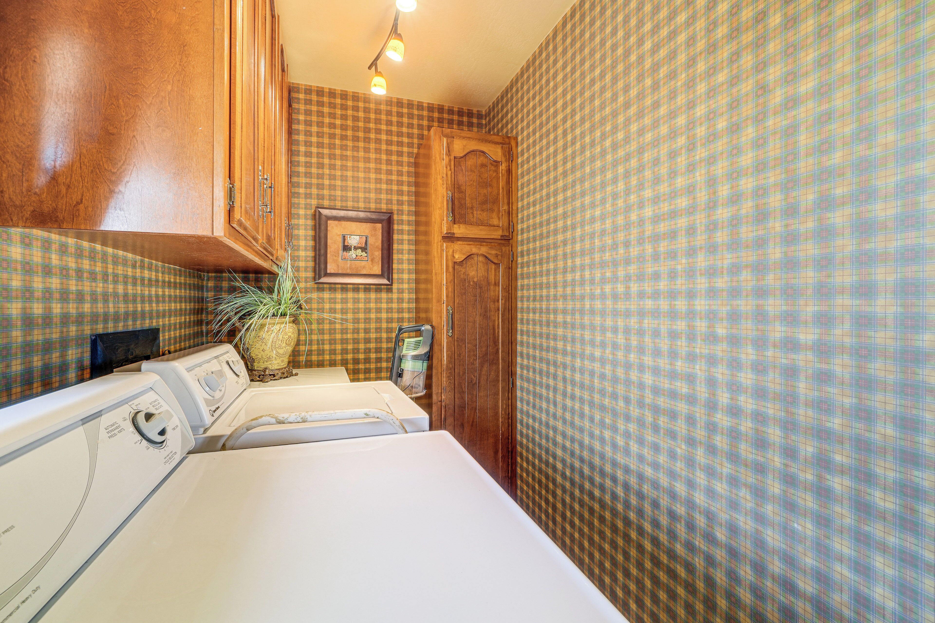 A laundry room with a washing machine, dryer, wooden cabinets, a framed picture, and a patterned wall.