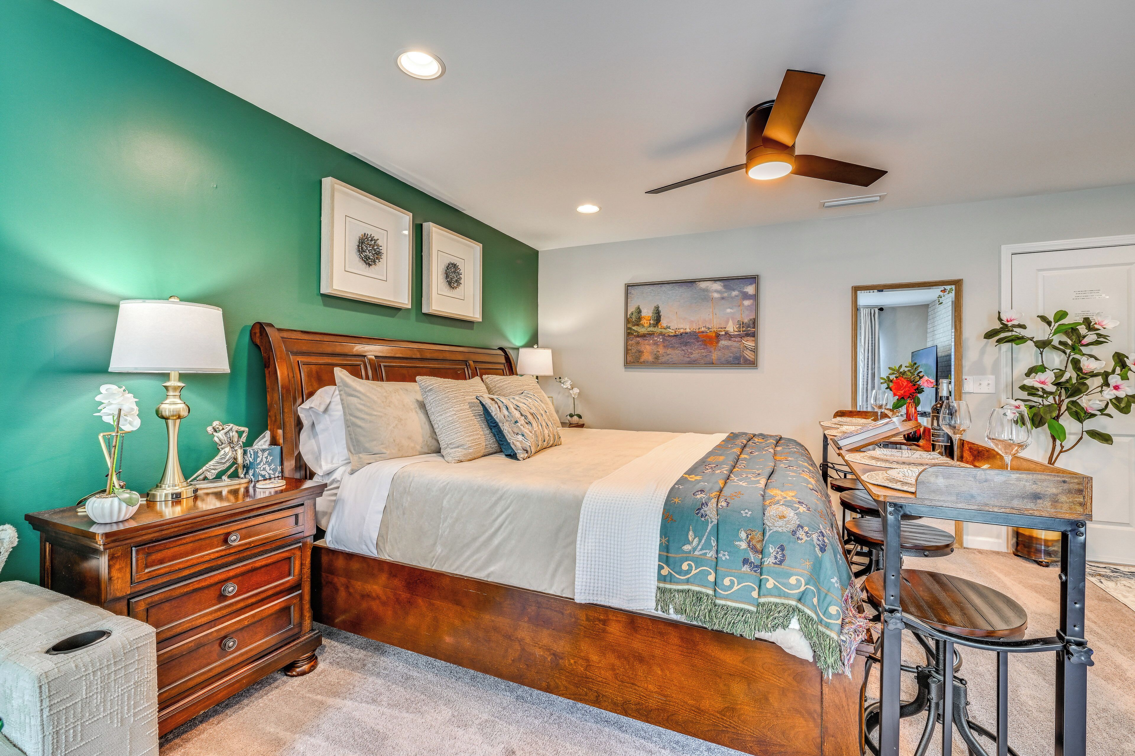 A bedroom with a bed, bedside tables, a dresser, a desk with chairs, a ceiling fan, and artwork on the walls.