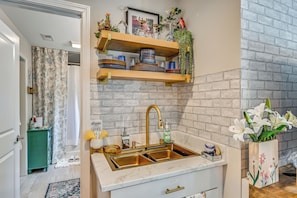 A kitchen with a double sink, a wooden shelf with dishes, a hanging plant, and a brick wall. - Deck, Patio! Charming Concord Studio Near Speedway (Concord)