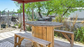 Une terrasse en bois avec une table ronde et des bancs, entourée d’une clôture en métal et d’arbres.