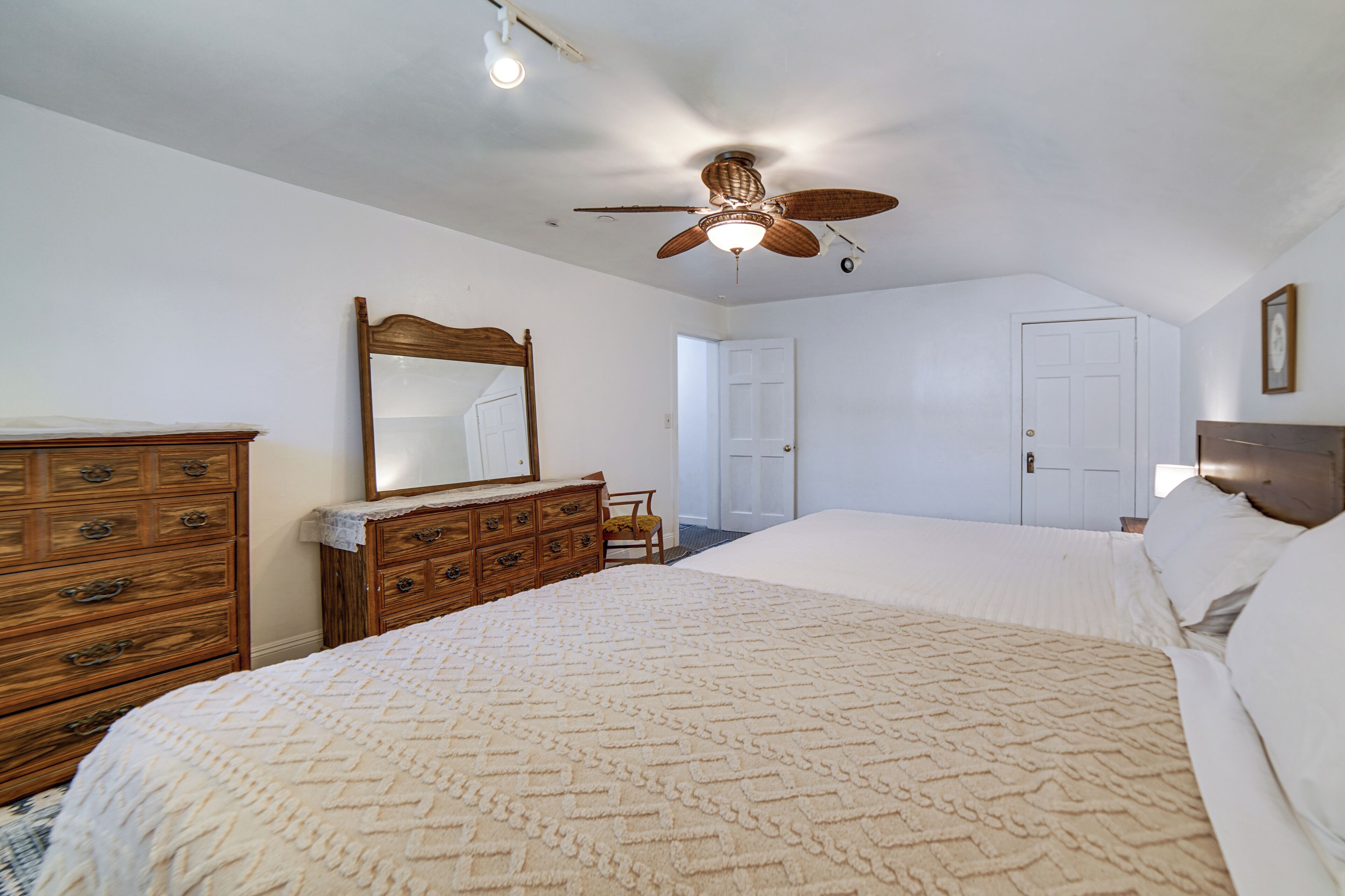 Quarto com uma cama grande, cómoda de madeira, ventoinha no teto e porta para outro espaço.