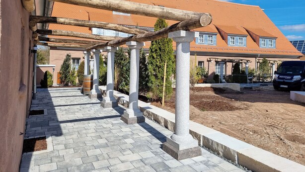 A covered outdoor area with stone columns, a paved walkway, and a building with a red roof in the background.