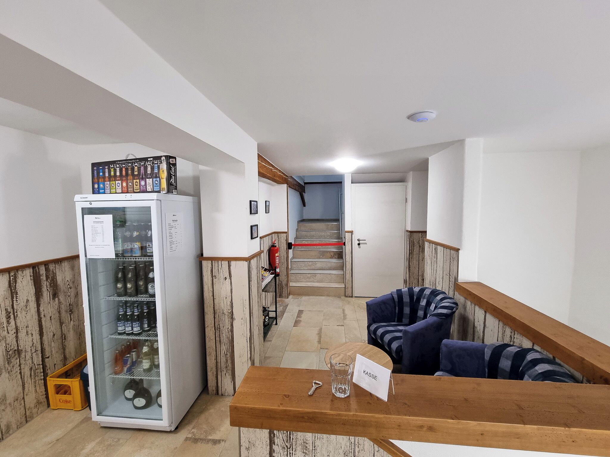 A reception area with a refrigerator stocked with beverages, a wooden counter with a name card, and a staircase in the background.