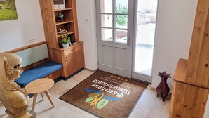 Un área de recepción con un escritorio de madera, un banco, una planta en maceta, una puerta con paneles de vidrio y un felpudo con texto.