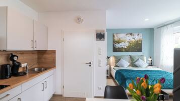Cozinha moderna com cafeteira, pia e armários. Há uma porta que dá para um quarto com cama, almofadas e mesa de cabeceira. É visível uma janela com cortinas, e um vaso com flores está sobre uma mesa.