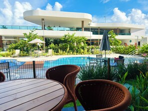 Un espace aménagé au bord de la piscine, avec des fauteuils en osier, une piscine carrelée et, en arrière-plan, un bâtiment moderne doté de balcons.