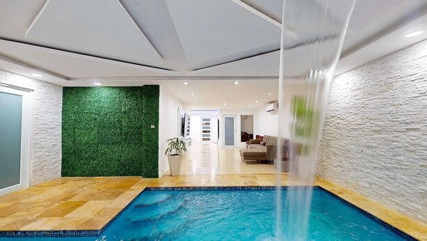 A modern indoor pool with a clear view of the adjacent living area, a green wall, and a stone-textured wall. - Modern 3BR Apt  AC + Comfort & Style (Trujillo Alto)