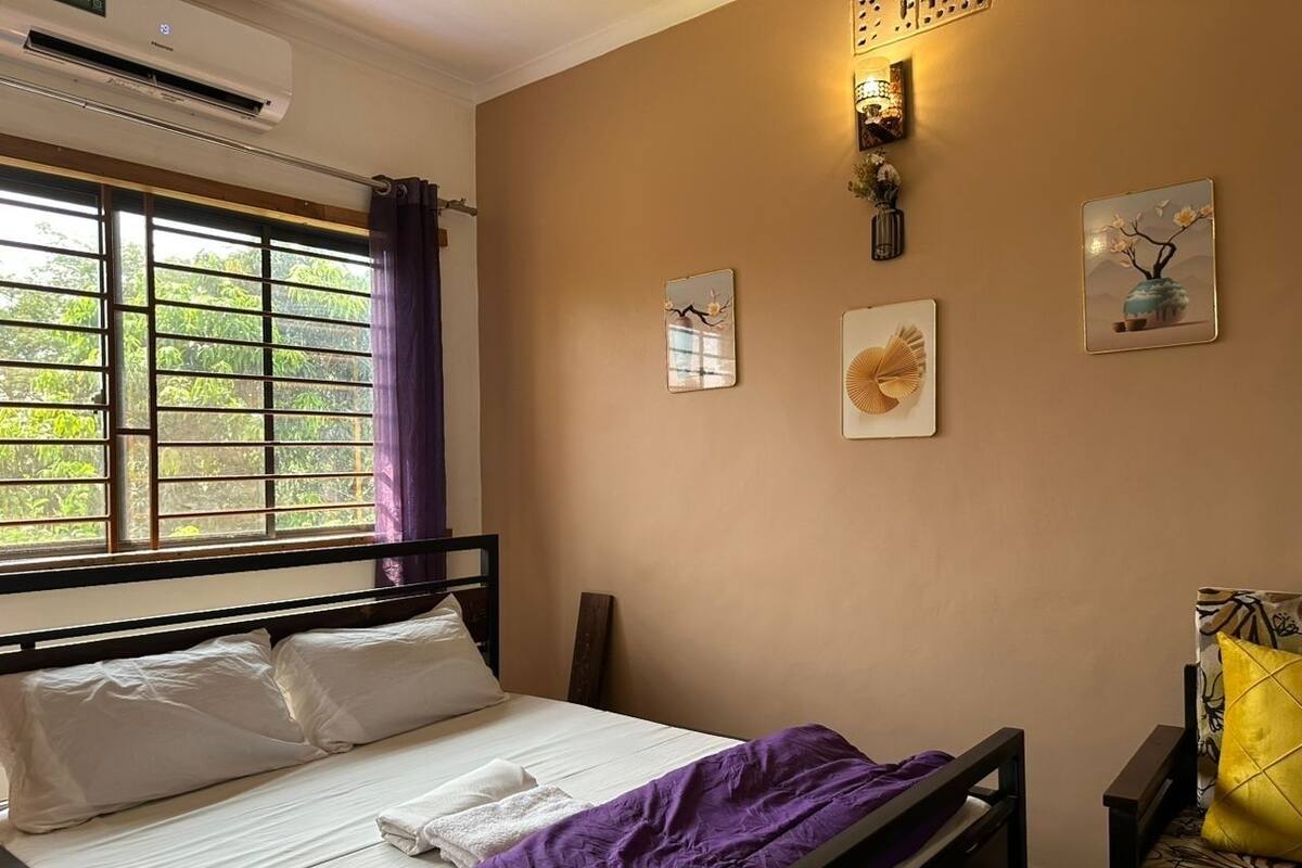 A bedroom with a bed, a window with curtains, a wall-mounted air conditioner, and a nightstand with a lamp.