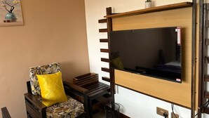 Une chambre avec un lit, un téléviseur fixé au mur, une chaise avec un coussin jaune et une petite plante sur l’étagère.