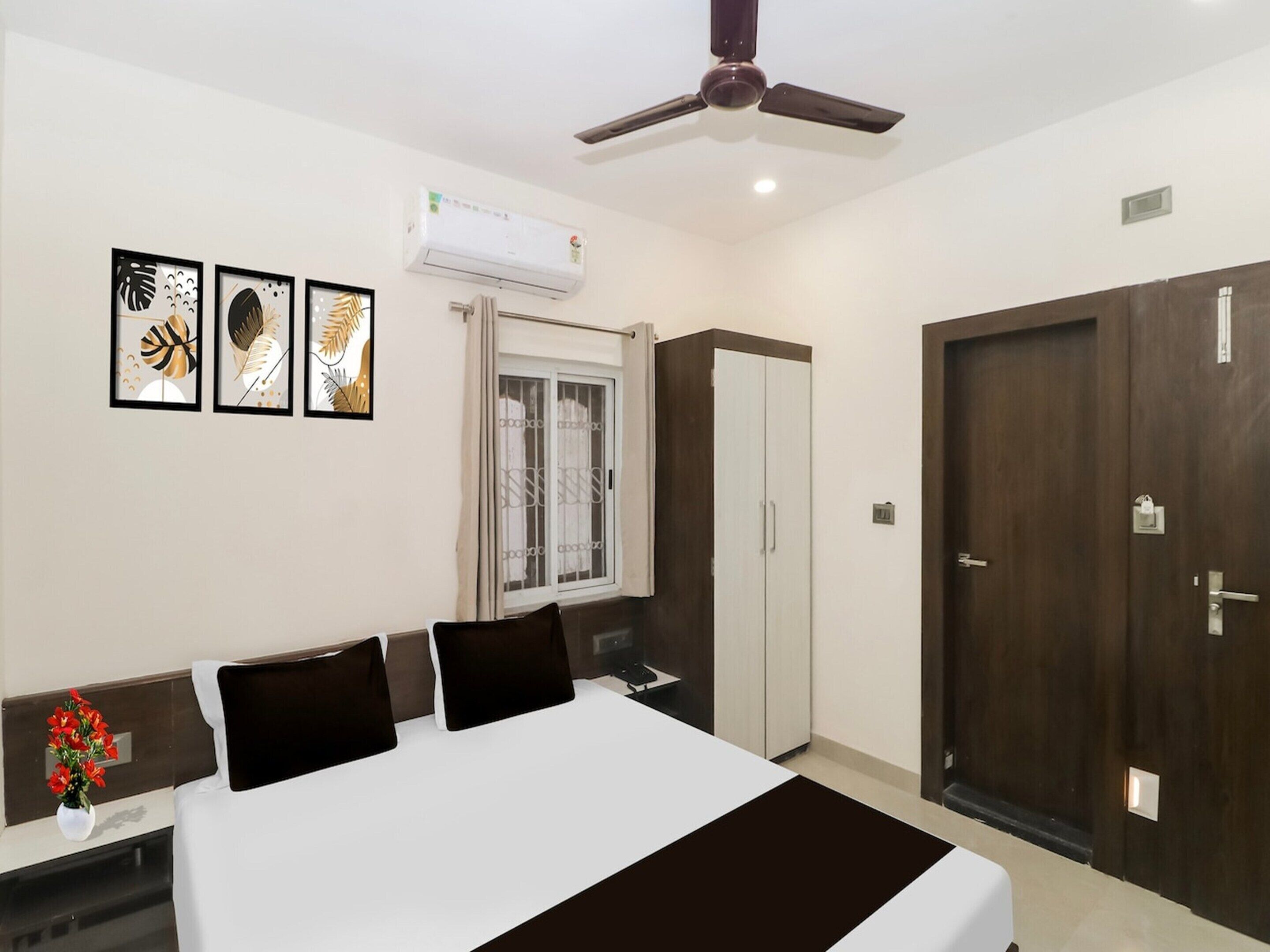 A hotel room with a bed, a wardrobe, a window with curtains, an air conditioning unit, and framed artwork on the wall.