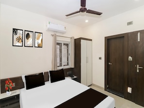 Una habitación de hotel con cama, armario, ventana con cortinas, aire acondicionado y cuadros en la pared.