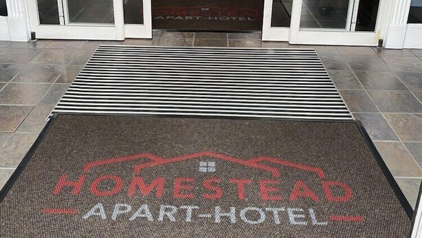 Ein Hotel Eingangsbereich mit Fußmatte mit der Aufschrift 'HOMESTEAD APART-HOTEL', Glastüren, einer Rezeption und einem Wartebereich.
