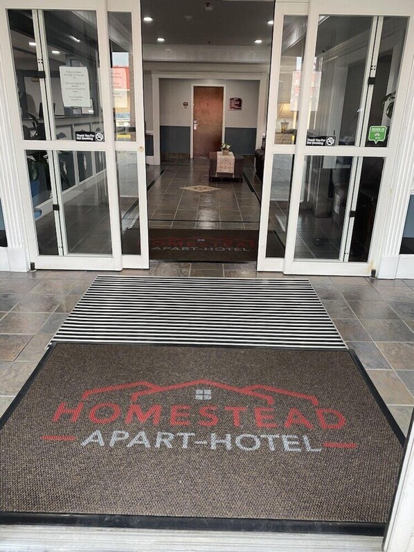 Entrada de hotel con felpudo que dice 'HOMESTEAD APART-HOTEL', puertas de cristal, mostrador de recepción y área de espera.