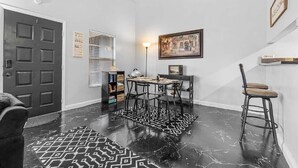 Un pasillo con una puerta oscura, un tapete estampado, una mesa con sillas, una lámpara y un cuadro enmarcado en la pared.