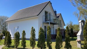 Une maison à deux étages avec un balcon, une porte d’entrée et un petit jardin planté de conifères.