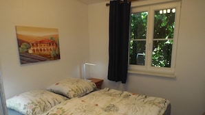 Ein Schlafzimmer mit einem Bett, einem Nachttisch, einer Lampe, einem Fenster mit Vorhängen und einem Bild an der Wand.