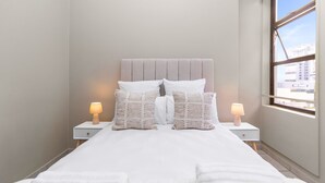 Ein ordentlich gemachtes Bett mit weißen Bettwäsche und Kissen, zwei Nachttische mit Lampen und ein Fenster mit Blick auf Gebäude.