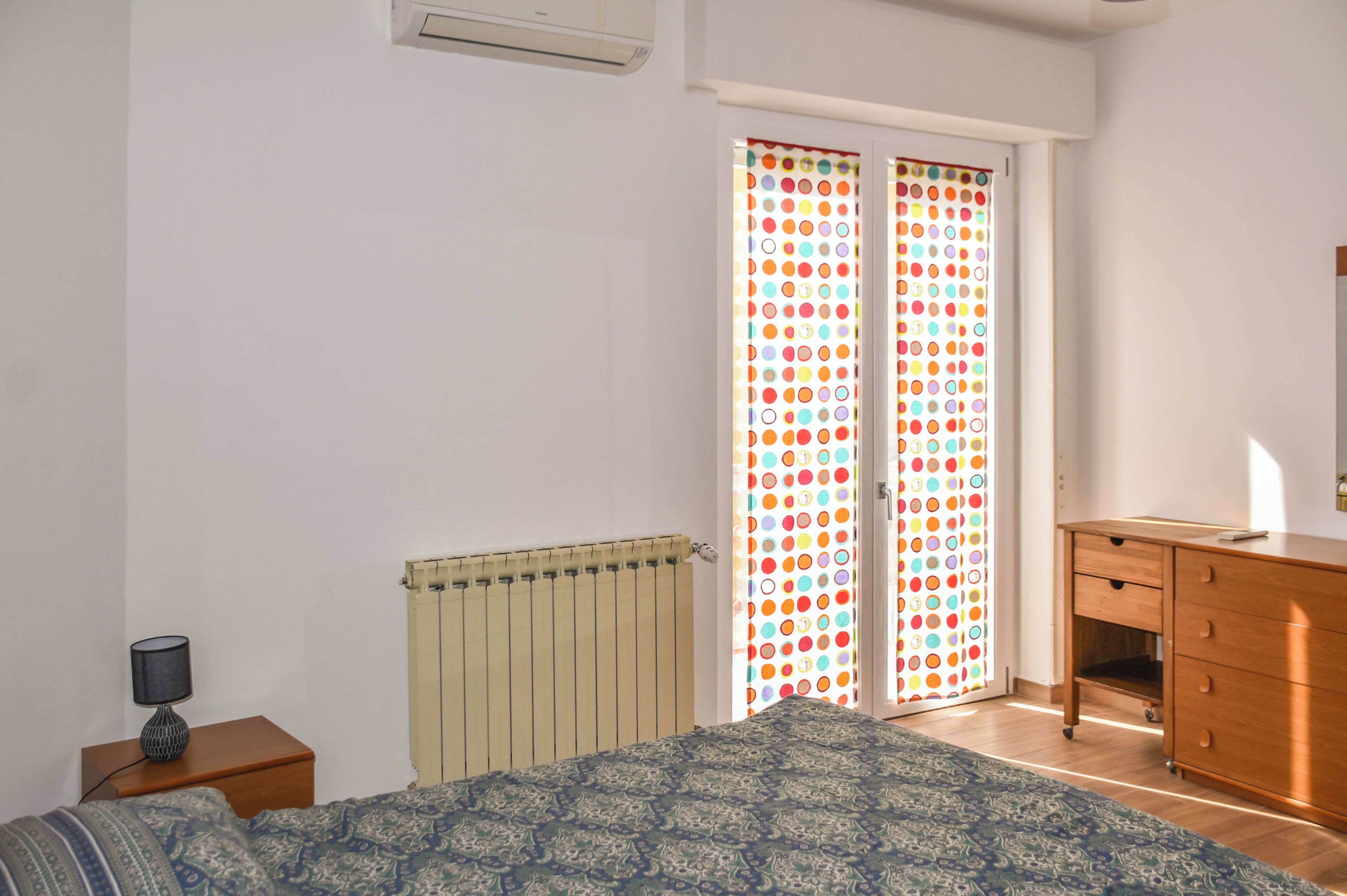 A bedroom with a bed, a nightstand, a dresser, a radiator, and a window with a colorful curtain.