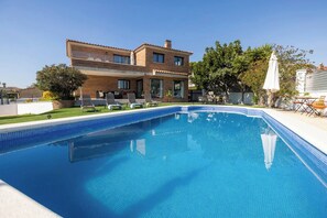 Une piscine rectangulaire dont les bords sont recouverts de carreaux, entourée d’une pelouse et bordée d’un bâtiment à deux étages doté d’un balcon.