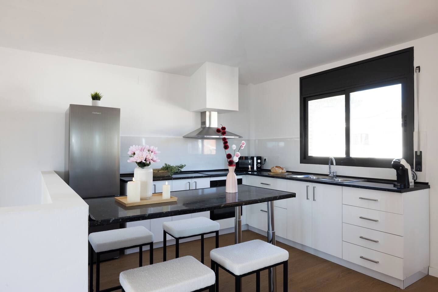 A modern kitchen with a central island, white cabinetry, a stainless steel range hood, and a window with a black frame.