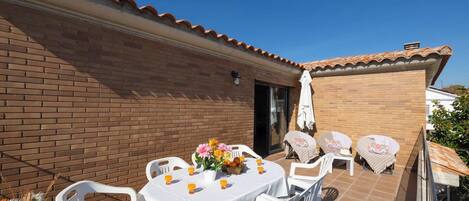 A patio with a round table set for four, surrounded by white chairs, a brick wall, and a tiled roof.