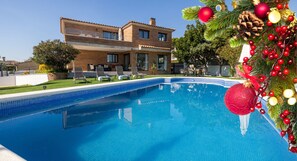 Una piscina con borde de azulejos, una casa de dos pisos al fondo y decoración navideña.