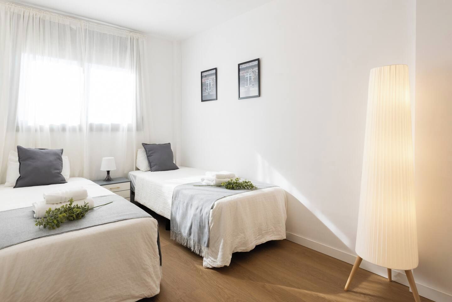 A hotel room with two beds, a bedside table with a lamp, and a large window with sheer curtains.