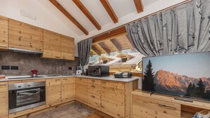 A modern kitchen with wooden cabinets, a built-in oven, a large window with curtains, and a television displaying a mountain landscape.