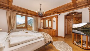 A bedroom with a bed, wooden ceiling, a chandelier, a television, and a view of mountains through the window.