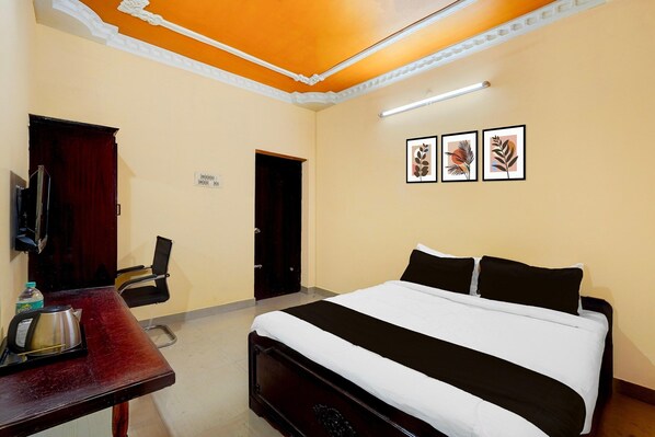 A hotel room with a bed, a desk with a kettle, a chair, and framed artwork on the wall. - Deluxe Sree Residency (Sriperumbudur)