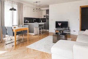 Ein modernes Wohnzimmer mit einer Couch, einem Esstisch mit Stühlen, einer Küchenzeile mit Kühlschrank und einem Fernseher.