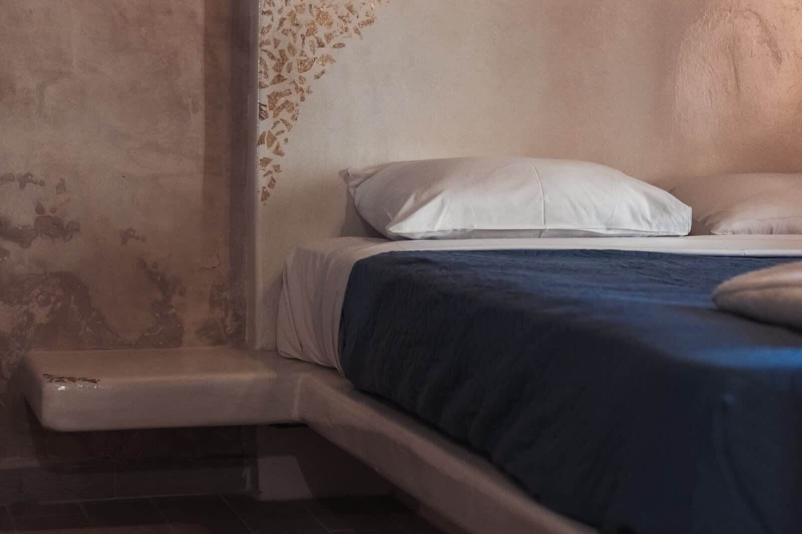 Un lit avec un oreiller blanc et une couverture bleue, une table de chevet blanche et un mur à texture.