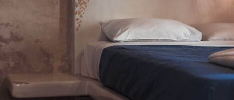 Un lit avec un oreiller blanc et une couverture bleue, une table de chevet blanche et un mur à texture.