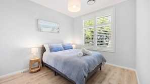 Een slaapkamer met een bed, een nachtkastje, een lamp, een raam met zonwering en een schilderij aan de muur.