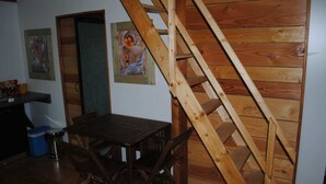 Una escalera de madera que sube a un nivel superior, una mesa de comedor con sillas y dos obras de arte enmarcadas en la pared.