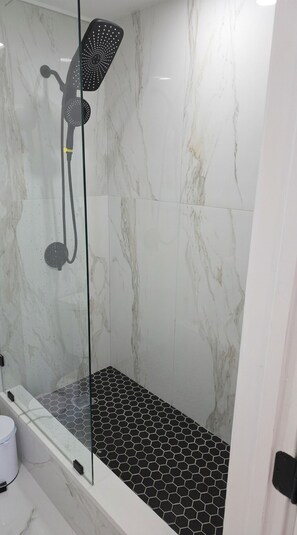 A glass-enclosed shower with a black and white hexagonal tile floor, marble walls, and a handheld showerhead.