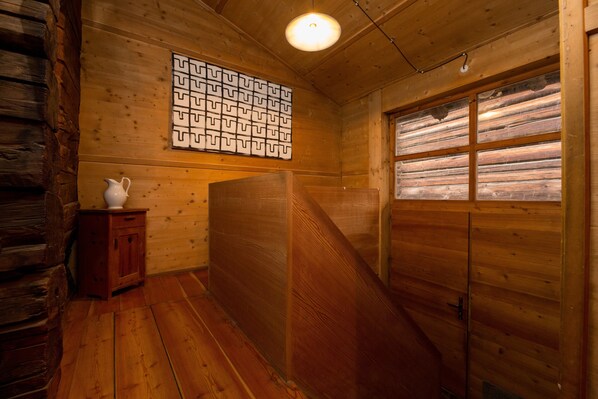 Interior de una cabaña de madera con piso de madera, una jarra blanca sobre un mueble pequeño, una lámpara de pared y una ventana con contraventanas de madera.
