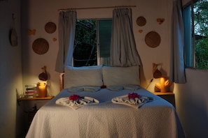 A neatly made bed with a white quilt, two pillows, and a decorative headboard. There are two bedside tables with lamps, a window with curtains, and wall decorations. - Praia do Sagi (Baía Formosa)