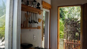 A kitchen with wooden shelves holding pots, pans, and utensils, a gas stove, and a view of a bamboo fence outside. - Praia do Sagi (Baía Formosa)