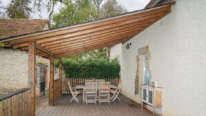 Une terrasse couverte avec une table et des chaises en bois, un petit meuble et un mur en pierre.