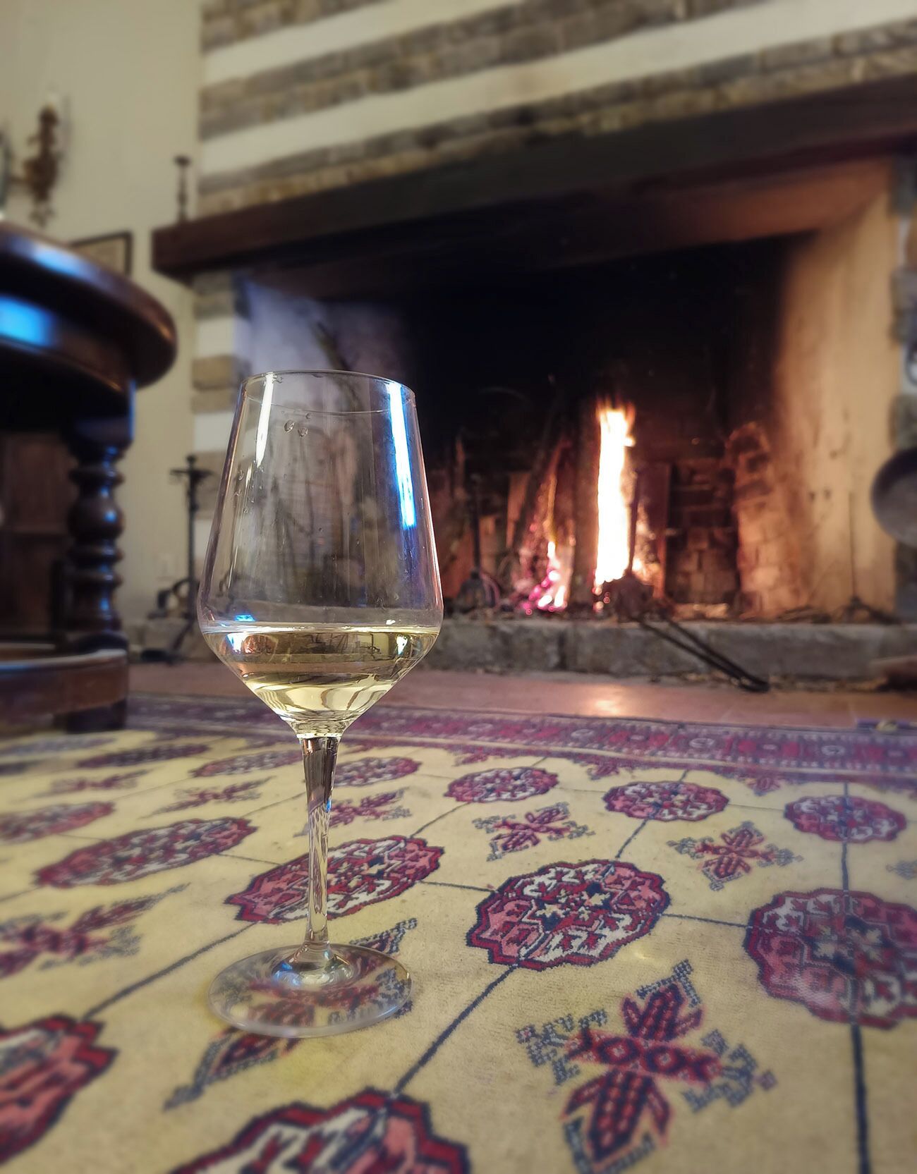 Ein Glas Weißwein auf einem gemusterten Teppich, im Hintergrund ein Kamin.