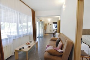 Ein Wohnzimmer mit einer Couch, einem Couchtisch und einem Fernseher. Im Hintergrund ist eine Küchenzeile zu sehen.