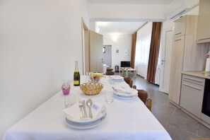Ein Essbereich mit Tisch für vier Personen, einem Fernseher im Hintergrund und einer Küchenzeile mit Schränken und einem Backofen.